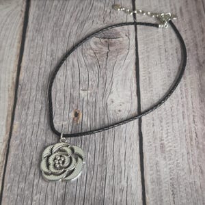 以下が含まれることがあります： 花柄のデザインが施されたシルバーカラーのペンダントネックレス。ペンダントは、シルバーカラーの留め金が付いた黒色の編み込みコードネックレスから吊り下げられています。ネックレスは木製の表面に表示されています。