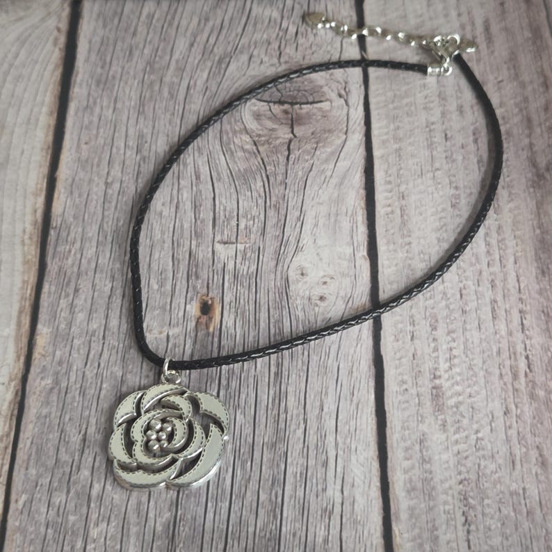 以下が含まれることがあります： 花柄のデザインが施されたシルバーカラーのペンダントネックレス。ペンダントは、シルバーカラーの留め金が付いた黒色の編み込みコードネックレスから吊り下げられています。ネックレスは木製の表面に表示されています。