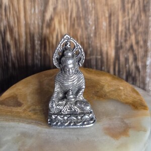 May include: A small silver statue of a seated Buddha with a decorative halo. The statue is on a light brown and white marble surface.