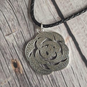 以下が含まれることがあります： ハート型のアクセントが付いた、詳細なフローラルデザインのシルバーペンダントネックレス。ペンダントは黒い編み込みコードから吊り下げられています。木製の表面に表示されています。