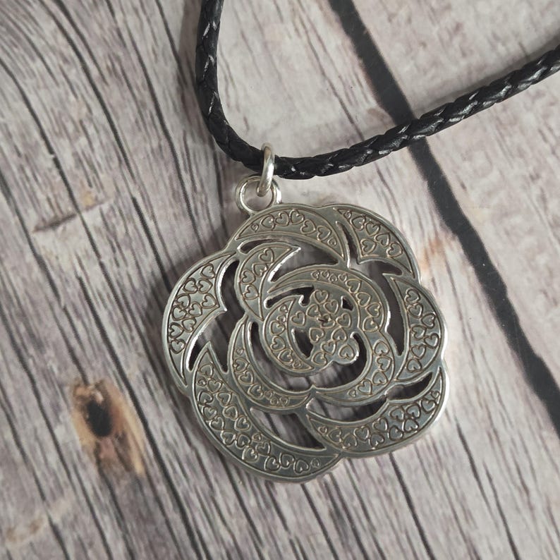 以下が含まれることがあります： ハート型のアクセントが付いた、詳細なフローラルデザインのシルバーペンダントネックレス。ペンダントは黒い編み込みコードから吊り下げられています。木製の表面に表示されています。