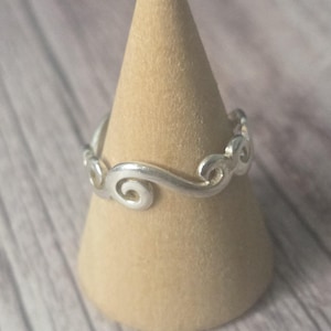 Puede incluir: Un anillo plateado con un diseño de nube arremolinada. El anillo se muestra sobre un soporte cónico de color beige. El diseño del anillo presenta una serie de formas de nubes estilizadas y conectadas.