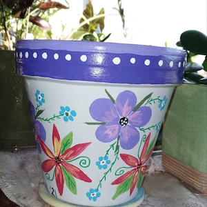 May include: A white terracotta flower pot with a purple rim and white polka dots. The pot is hand-painted with colorful flowers and green leaves. The pot is sitting on a white saucer.
