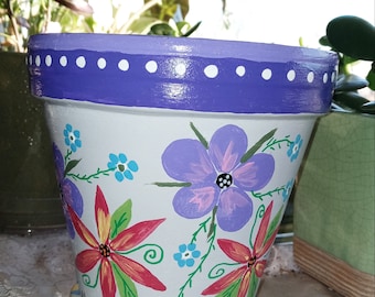 May include: A white terracotta flower pot with a purple rim and white polka dots. The pot is hand-painted with colorful flowers and leaves. The pot is sitting on a white saucer.