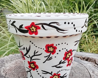 May include: White terracotta flower pot with black and red floral design. The pot is decorated with black branches, red flowers with yellow centers, and black dots. The bottom of the pot is signed "AL '21".