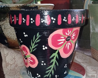 May include: Black terracotta flower pot with a red and white border, painted with pink flowers and green leaves. The pot is sitting on a blue and white saucer with the text "AL 23" on the bottom.