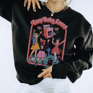 May include: Black sweatshirt with a graphic print of three girls in front of a tombstone and an oven with pies flying out. The text on the sweatshirt says "Easy-Bake Coven".