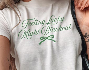May include: White t-shirt with the text "Feeling Lucky, Might Blackout" in green script. A green bow graphic is below the text. The shirt is paired with blue jeans.