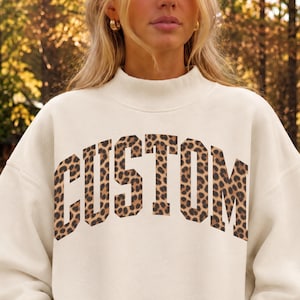 May include: An off-white sweatshirt with a mock neck and long sleeves, featuring the word CUSTOM printed across the chest in a bold, arched collegiate font with a brown and black leopard print pattern throughout the letters.