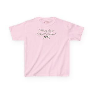 May include: A light pink t-shirt with the text "Feeling Lucky, Might Blackout" in a script font. A small green bow graphic is below the text. The brand name "The Haze Designs" is above the text.