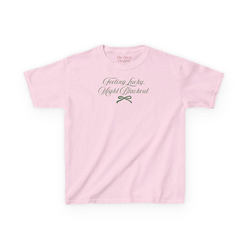 May include: A light pink t-shirt with the text "Feeling Lucky, Might Blackout" in a script font. A small green bow graphic is below the text. The brand name "The Haze Designs" is above the text.
