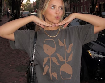 May include: Dark gray t-shirt with a brown graphic of a wine glass and a vase with leaves. The shirt is paired with blue jeans and a gold necklace. A black purse hangs from the shoulder.