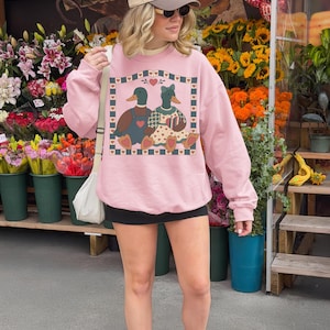 May include: Pink sweatshirt featuring a graphic of two ducks with hearts. The ducks are wearing overalls and a dress. The sweatshirt is worn by a person in front of a flower stand.