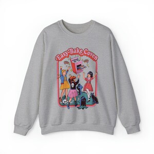 May include: A gray sweatshirt with a graphic print of three girls in front of an Easy-Bake Oven. The text "Easy-Bake Coven" is written in a pink and red font above the oven. The girls are wearing colorful dresses and are holding up pies. A black cat is sitting in the background.