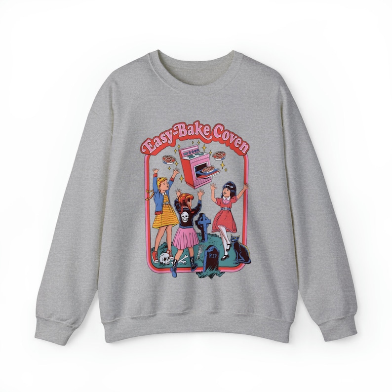 May include: A gray sweatshirt with a graphic print of three girls in front of an Easy-Bake Oven. The text "Easy-Bake Coven" is written in a pink and red font above the oven. The girls are wearing colorful dresses and are holding up pies. A black cat is sitting in the background.