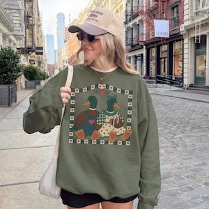 May include: An olive green sweatshirt featuring a charming illustration of two ducks, one male and one female, within a heart-bordered frame. The ducks are adorned with clothing and accessories. The sweatshirt is worn by a person.