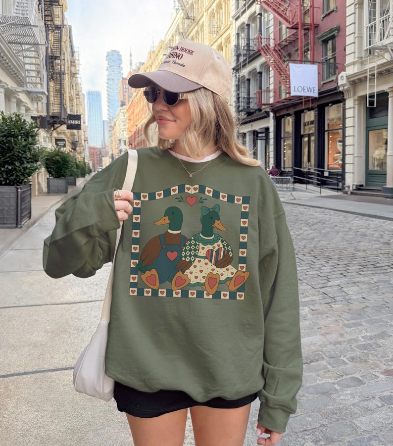May include: An olive green sweatshirt featuring a charming illustration of two ducks, one male and one female, within a heart-bordered frame. The ducks are adorned with clothing and accessories. The sweatshirt is worn by a person.
