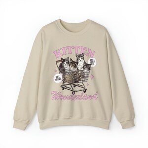 Puede incluir: Sudadera beige con un estampado gr&aacute;fico de cuatro gatitos en un carro de la compra. El texto "Kitten Wonderland" est&aacute; impreso debajo de la imagen. Los textos "99% Meows" y "100% Cute" est&aacute;n impresos en el gr&aacute;fico.