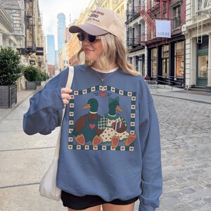 May include: A blue sweatshirt featuring a graphic of two ducks in love, surrounded by hearts. The ducks are wearing outfits and are set against a heart-filled border. The person in the image is wearing a baseball cap and sunglasses.