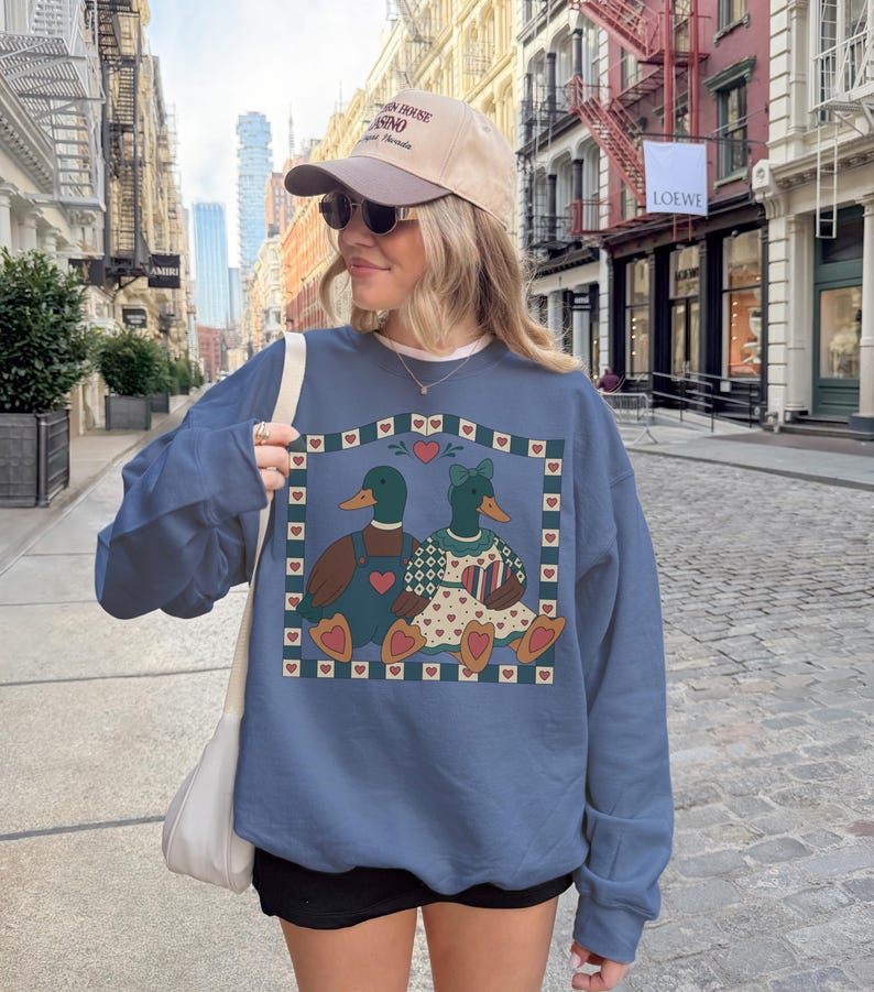 May include: A blue sweatshirt featuring a graphic of two ducks in love, surrounded by hearts. The ducks are wearing outfits and are set against a heart-filled border. The person in the image is wearing a baseball cap and sunglasses.