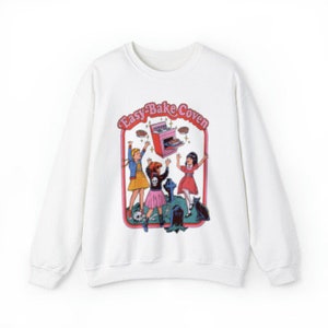May include: White sweatshirt with a graphic print of three girls in front of an Easy-Bake Oven. The text "Easy-Bake Coven" is printed above the oven.