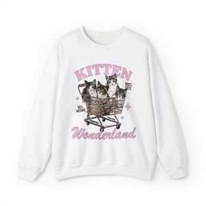 Puede incluir: Sudadera blanca con un estampado gr&aacute;fico rosa de cuatro gatitos en un carrito de la compra. El texto del gr&aacute;fico dice "Kitten Wonderland" y "99% Meows" y "100% Cute".