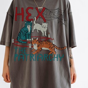 Puede incluir: Camiseta gris oscuro con un gr&aacute;fico de tres gatos, velas y una telara&ntilde;a. El texto dice "HEX" en rojo y "THE PATRIARCHY" en turquesa. La camiseta tiene mangas cortas y un corte holgado.