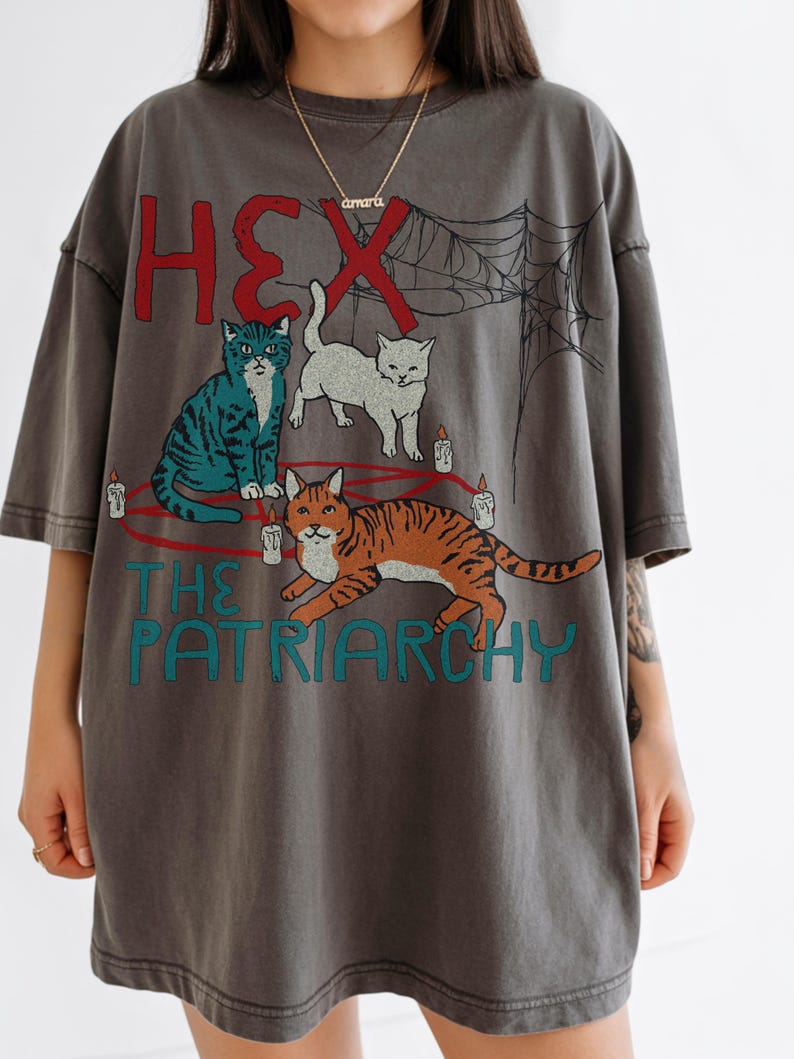 Puede incluir: Camiseta gris oscuro con un gr&aacute;fico de tres gatos, velas y una telara&ntilde;a. El texto dice "HEX" en rojo y "THE PATRIARCHY" en turquesa. La camiseta tiene mangas cortas y un corte holgado.