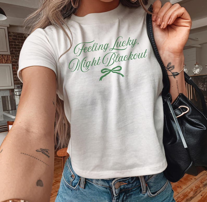 White t-shirt with the text "Feeling Lucky, Might Blackout" in green script. A green bow graphic is below the text. The shirt is paired with blue jeans.
