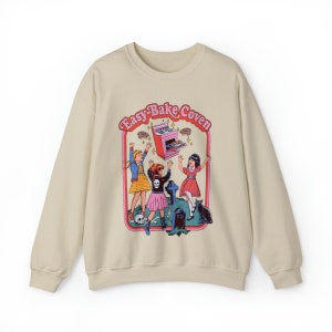 May include: A beige sweatshirt with a graphic print of three girls in front of a tombstone. The graphic features a pink Easy-Bake Oven with the text "Easy-Bake Coven".