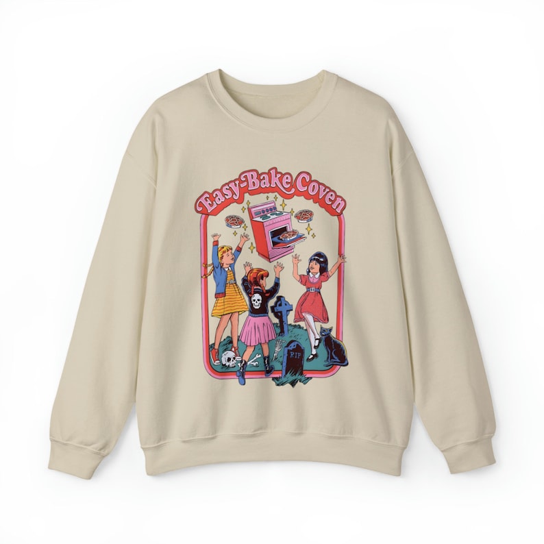 May include: A beige sweatshirt with a graphic print of three girls in front of a tombstone. The graphic features a pink Easy-Bake Oven with the text "Easy-Bake Coven".