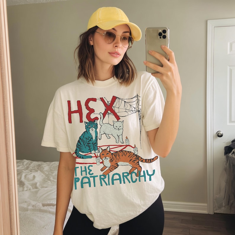 Person wearing a white oversized graphic T-shirt with bold red text that reads HEX above an illustrated design of a cat casting a spell with the words ‘HEX THE PATRIARCHY’ in teal. The shirt has a fun, feminist, vintage-inspired style. Model is taking a mirror selfie, styled with a yellow baseball cap and sunglasses.