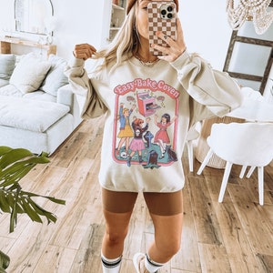 May include: A beige sweatshirt with a graphic print of three girls in front of an Easy-Bake Oven. The text "Easy-Bake Coven" is printed on the sweatshirt.