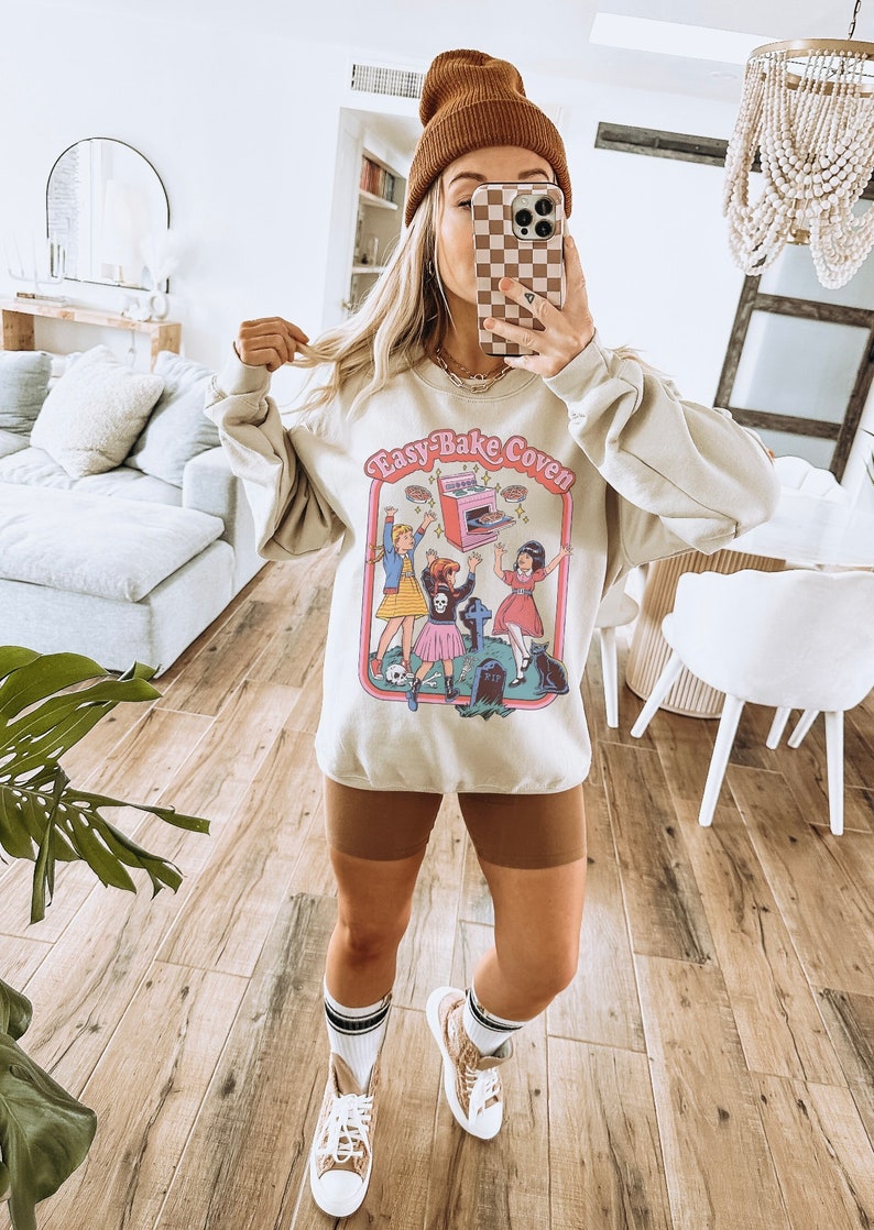 May include: A beige sweatshirt with a graphic print of three girls in front of an Easy-Bake Oven. The text "Easy-Bake Coven" is printed on the sweatshirt.