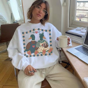 White sweatshirt featuring a graphic of two ducks in a heart-bordered frame. One duck wears overalls, the other a dress. The design includes hearts and a floral pattern. The sweatshirt has long sleeves.