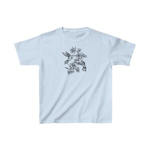 K&ouml;nnte beinhalten: Ein hellblaues T-Shirt mit einer schwarz-wei&szlig;en Illustration von zwei Kolibris im Flug, die sich gegen&uuml;berstehen und sich mit ihren Schn&auml;beln ber&uuml;hren. Die Kolibris sind von schwarzen und wei&szlig;en Blumen umgeben.