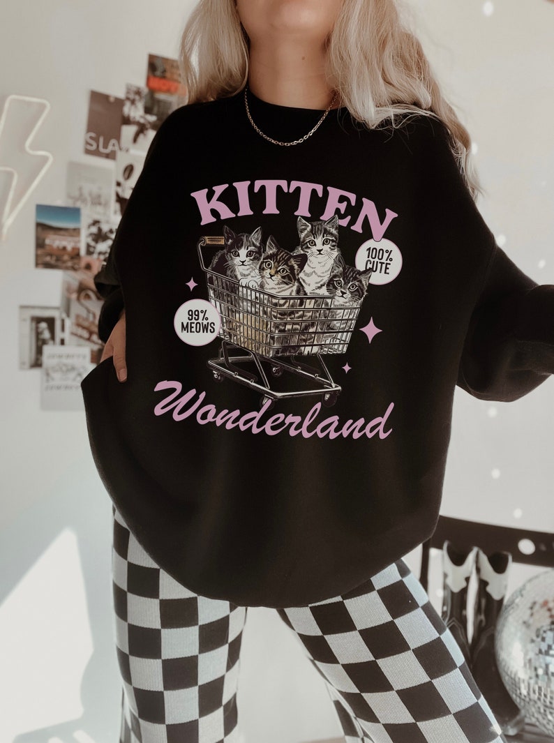 Puede incluir: Sudadera negra con un gr&aacute;fico rosa de gatitos en un carrito de compras. El gr&aacute;fico dice "Kitten Wonderland" con el texto "99% Meows" y "100% Cute" en el carrito.