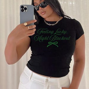 May include: A black t-shirt with the text "Feeling Lucky, Might Blackout" in green script. The shirt is paired with white pants and a gold chain necklace. The person is wearing sunglasses and holding a phone.