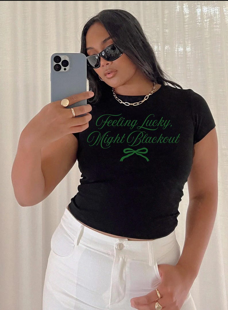 May include: A black t-shirt with the text "Feeling Lucky, Might Blackout" in green script. The shirt is paired with white pants and a gold chain necklace. The person is wearing sunglasses and holding a phone.