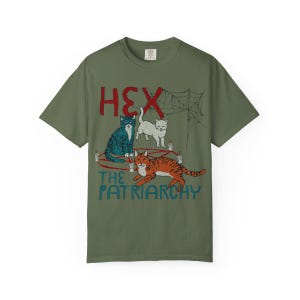 Puede incluir: Camiseta verde oliva con un dise&ntilde;o gr&aacute;fico que presenta tres gatos, una telara&ntilde;a y el texto "HEX THE PATRIARCHY" en rojo y azul. Los gatos est&aacute;n en varios colores y poses, con velas y un dise&ntilde;o de pentagrama.