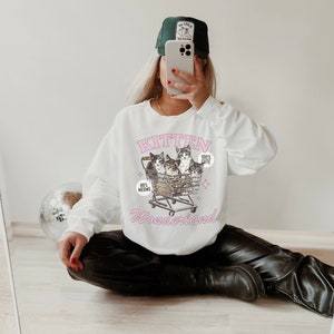 Puede incluir: Sudadera blanca con un gr&aacute;fico rosa de gatitos en un carrito de compras. El gr&aacute;fico dice "Kitten Wonderland" y "99% Meows" y "100% Cute".