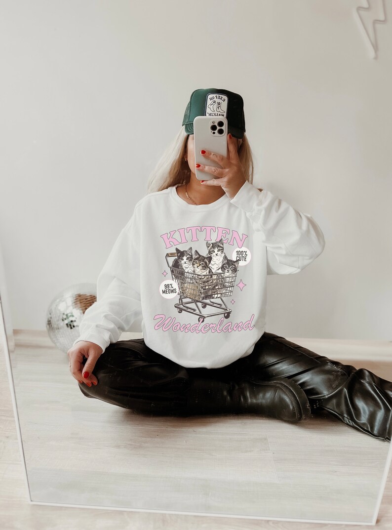 Puede incluir: Sudadera blanca con un gr&aacute;fico rosa de gatitos en un carrito de compras. El gr&aacute;fico dice "Kitten Wonderland" y "99% Meows" y "100% Cute".