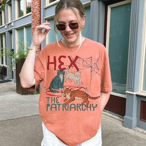 Puede incluir: Camiseta naranja con el texto "HEX THE PATRIARCHY" y un gr&aacute;fico de tres gatos, velas y una telara&ntilde;a. La camiseta es de corte holgado y tiene mangas cortas.