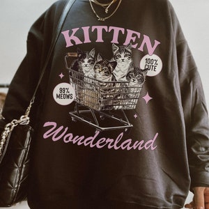 Puede incluir: Una sudadera gris oscuro con un gr&aacute;fico rosa de gatitos en un carrito de compras. El gr&aacute;fico dice "Kitten Wonderland" y "99% Meows" y "100% Cute".