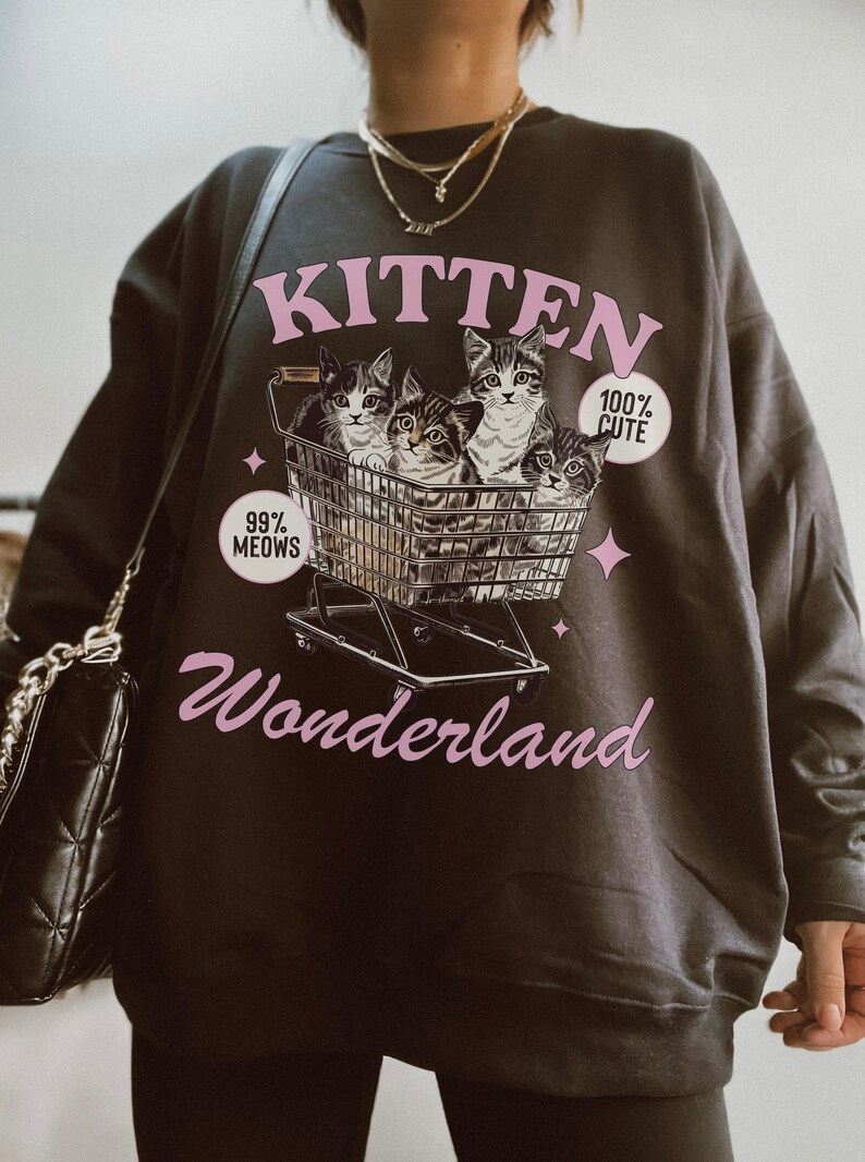 Puede incluir: Una sudadera gris oscuro con un gr&aacute;fico rosa de gatitos en un carrito de compras. El gr&aacute;fico dice "Kitten Wonderland" y "99% Meows" y "100% Cute".