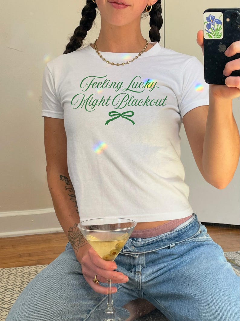 May include: White t-shirt with the text "Feeling Lucky, Might Blackout" in green script. The shirt has a crew neck and short sleeves. The person is wearing blue jeans and holding a martini glass with an olive.