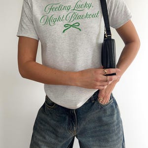 May include: A light gray t-shirt with the text "Feeling Lucky, Might Blackout" in green script and a green bow graphic. The person is wearing blue jeans and carrying a black purse.