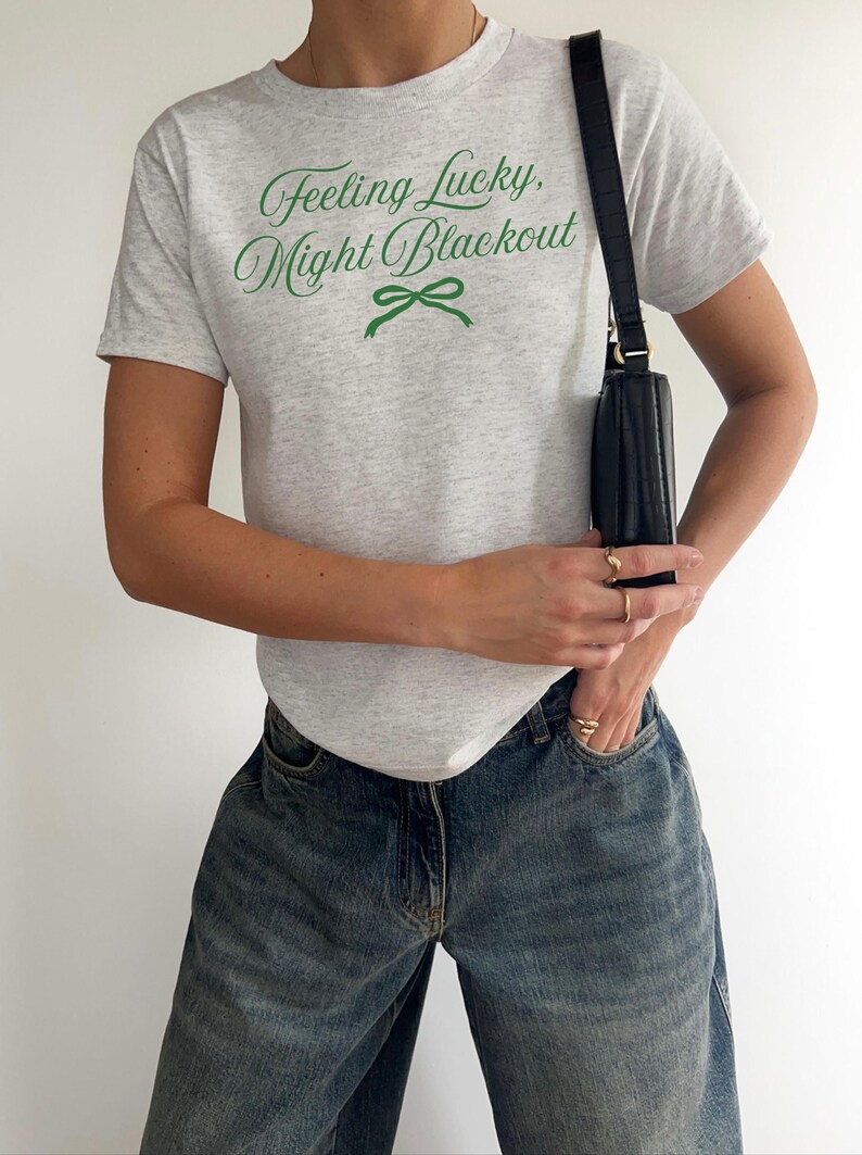 May include: A light gray t-shirt with the text "Feeling Lucky, Might Blackout" in green script and a green bow graphic. The person is wearing blue jeans and carrying a black purse.