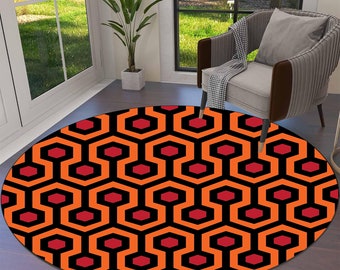 Può includere: Un tappeto rotondo con un motivo geometrico ripetuto in arancione, nero e rosso. Il design presenta esagoni interconnessi e forme rosse più piccole, creando un effetto visivo audace. Il tappeto è posato su un pavimento in legno.