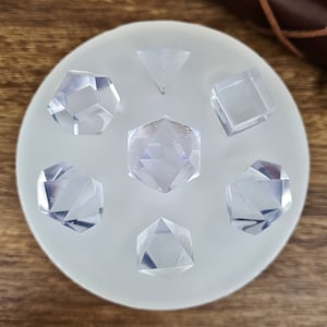 May include: A silicone mould for making clear resin dice. The mould has seven cavities, each shaped like a different polyhedron. The mould is on a wooden surface.